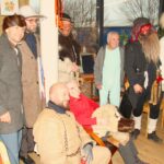 Skupina oseb v pustnih kostumih stoji skupaj v prostoru, ena oseba sedi na stolu in drži kos krzna.