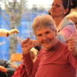 Ženska dvigne roke med plesom, v ozadju druge osebe sodelujejo pri dejavnosti.