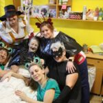 Več oseb v pustnih kostumih pozira ob postelji v sobi.