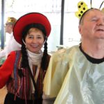 Ženska v tradicionalnem kostumu stoji ob moškem z naglavnim obročem z antenami v prostoru z drugimi osebami v ozadju.