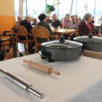 Na mizi s prtom sta dva lonca s pokrovom in več valjarjev, v ozadju pa osebe sedijo za mizami v skupnem prostoru.