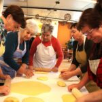 Več žensk stoji okoli mize in z valjarji razvaljuje testo na pomokani površini.