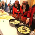 Oseba s penovko obrača koščke testa, ki se cvrejo v vročem olju v dveh posodah na mizi, druge osebe stojijo ob strani in opazujejo.