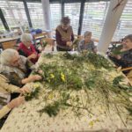 Več oseb sedi okoli mize in iz vej ter cvetja izdeluje aranžmaje.