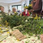 Na mizi so razporejene veje, listje in vrvica, okoli pa osebe izdelujejo cvetlične aranžmaje.