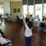 Starejši sedijo ob mizah in z dvignjenimi rokami sodelujejo pri skupinski vadbi, ki jo vodi oseba spredaj.