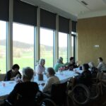 Skupina starejših sedi ob dolgi mizi ob oknih in ustvarja z različnimi materiali.