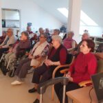 Skupina oseb sedi na stolih in invalidskih vozičkih v prostoru ter gleda proti prenosnemu računalniku na mizi.