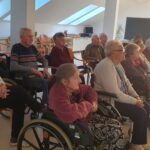 Osebe sedijo v vrsti na stolih in invalidskih vozičkih ter spremljajo projekcijo v prostoru z velikimi strešnimi okni.