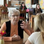 Dve osebi sedita za mizo in se pogovarjata v prostoru z več mizami in stoli.