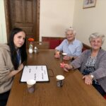 Tri osebe sedijo za mizo z lončki in papirnato rožo, na mizi je tabla za pisanje.