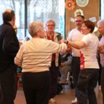 Več oseb stoji v krogu in se drži za roke med plesom v prostoru, okrašenem z baloni.