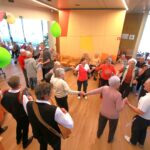 Skupina oseb pleše v krogu, medtem ko glasbeniki igrajo na instrumente ob robu prostora.