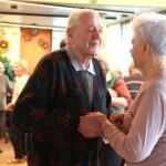 Moški in ženska stojita drug nasproti drugega in plešeta, v ozadju pa druge osebe opazujejo ali plešejo.