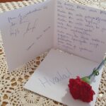 Odprta voščilnica z napisom »Hvala« in rokopisnim besedilom leži na mizi poleg rdečega nageljna.