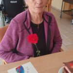 Ženska sedi za mizo in drži rdeč nagelj, na mizi pred njo sta kos torte in voščilnica z narisanimi cvetovi.