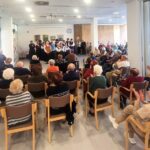 V večnamenskem prostoru starejši stanovalci sedijo na stolih in z aplavzom spremljajo nastop folklorne skupine v ospredju.