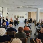 Folklorna skupina v tradicionalnih oblačilih nastopa pred občinstvom starejših, nekateri igrajo na glasbila, drugi pojejo.