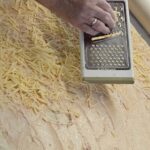 Oseba z ročnim strgalom riba sir po razvaljanem testu, premazanem z nadevom, na mizi s prtom.