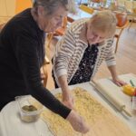 Dve ženski stojita ob mizi in posipata nariban sir ter začimbe po razvaljanem testu.