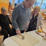 Moški z rezalnikom za testo reže razvaljano testo na trakove na mizi, ob njem stoji ženska, v ozadju pa druga oseba sedi na stolu.