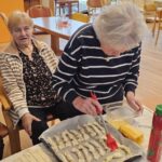 Ženska s kuhinjskim čopičem premaže kose testa na pekaču, medtem ko druga ženska sedi ob mizi v prostoru z velikimi okni.