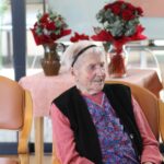 Udeleženka sedi in spremlja dogajanje, v ozadju pa lepo aranžirani šopki, ki še dodatno polepšajo prostor.