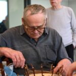 Stanovalec zbrano in natančno plete košarico, medtem ko v ozadju drugi sodelujejo pri dejavnosti.