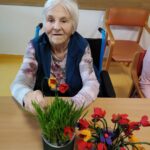 Starejša ženska sedi za mizo in drži majhen cvetlični aranžma, pred njo so še drugi izdelani cvetovi.