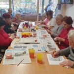 Skupina starejših oseb sedi za mizo in ustvarja; na mizi so barve, papir in različni materiali za izdelavo okraskov.