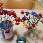 Na mizi sta dve posodi z barvitimi ročno izdelanimi cvetovi iz papirja ali podobnega materiala.