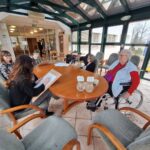 Več oseb sedi okoli mize v svetlem prostoru in riše na papir, medtem ko druge osebe opazujejo.