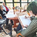 Oseba riše na papir, medtem ko dve osebi sedita nasproti in opazujeta dogajanje.