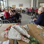 V ospredju je šopek rdečih nageljnov na mizi, v ozadju skupina oseb sedi v prostoru in posluša osebo, ki govori.