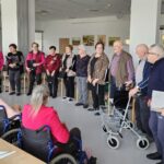 Skupina oseb stoji v vrsti in drži mape ali zvezke, pred njimi sedijo osebe na stolih in invalidskih vozičkih.