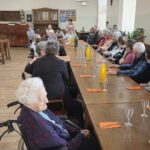 Starejši stanovalci sedijo za dolgo mizo, pripravljeno za praznovanje, medtem ko v ozadju otroci sodelujejo v programu.