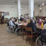 Skupina stanovalcev sedi za mizo, v ozadju pa otrok nastopa in zabava prisotne.