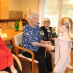 Otrok stoji pred sedečo osebo in ji izroča cvet, medtem ko druge osebe sedijo v ozadju.