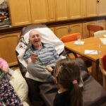 Otrok podaja cvet osebi, ki sedi v naslanjaču, medtem ko sta v bližini še dve osebi.