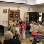 Skupina otrok se giblje po prostoru, medtem ko osebe sedijo ob robu in jih opazujejo.