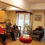 Več oseb sedi v prostoru okoli mize, na kateri je torta, in gleda proti sredini prostora.