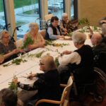 Več oseb sedi okoli mize ob oknu in iz vej ter zelenja izdeluje šopke.