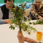 Oseba v ospredju drži šopek iz zelenja, medtem ko druge osebe sedijo za mizo in pripravljajo rastline.