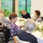 Več oseb sedi za mizo in razporeja veje ter zelenje, medtem ko sodelujejo v skupni dejavnosti.