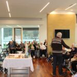 Oseba stoji pred skupino sedečih oseb in jim izroča zelenje, medtem ko je na mizi pred njimi postavljena dekoracija.