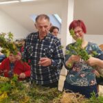 Več oseb stoji ob mizi in iz vej ter rastlin izdeluje šopke.