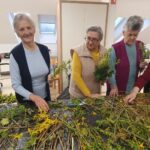 Tri osebe stojijo ob mizi, na kateri so razporejene veje, in izdelujejo šopke iz rastlin.