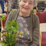 Oseba sedi na stolu in v rokah drži šopek zelenja in cvetja, medtem ko so druge osebe v ozadju.