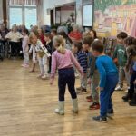 Skupina otrok stoji v prostoru in se giblje v vrsti, medtem ko osebe sedijo ob robu prostora in opazujejo.