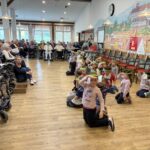 Otroci sedijo in klečijo na tleh v prostoru, medtem ko jih skupina oseb, sedečih ob strani, opazuje.